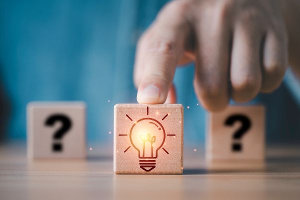 Geschäftsmann schiebt Holzblöcke, wobei ein Block mit einer leuchtenden Glühbirne vor einem Fragezeichen steht, um Kreativität, Innovation, Lösungen und Ideengenerierung im Business und bei der Problemlösung darzustellen
